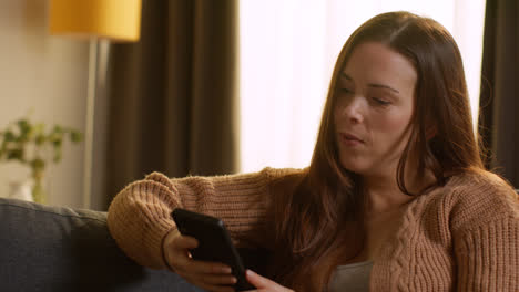 Woman-Sitting-On-Sofa-At-Home-Using-Mobile-Phone-To-Check-Social-Media-Message-And-Scrolling-Online-13